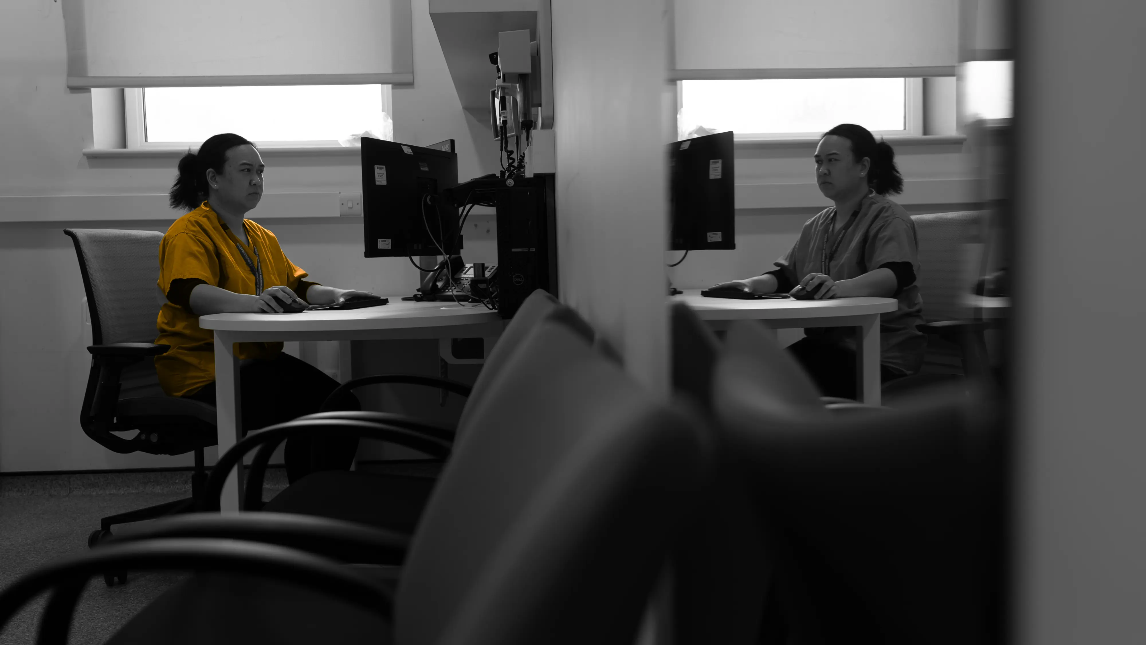Appointments at HIYOS. A female clinician sitting at a computer, with her reflection in a mirror. In a muted colour office setting, one wearing a yellow top and her reflection in a grey outfit.
