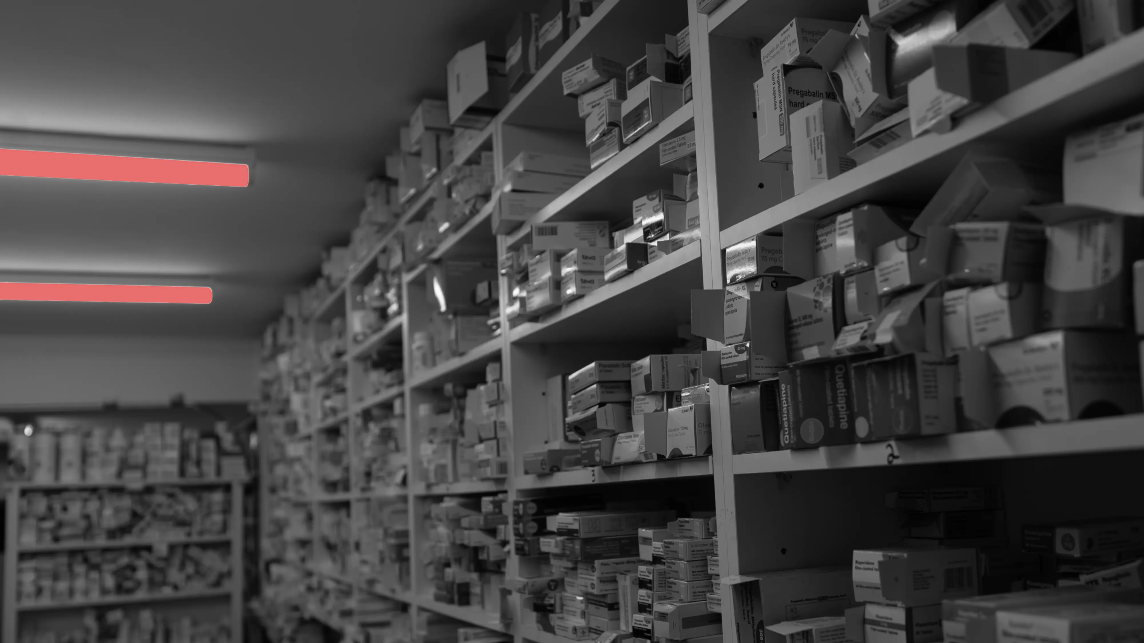 Medications in a pharmacy. A dimly lit pharmacy shelving unit filled with numerous prescription boxes, with two red overhead light fixtures illuminating the space.